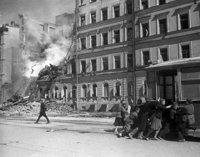Жители блокадного Ленинграда толкают трамвайный вагон подальше от фасада разрушенного бомбардировкой дома.